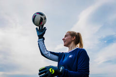 A girl with blond hair is wearing the navy blue training top with long sleeves, football gloves and is balancing a ball on her right hand.