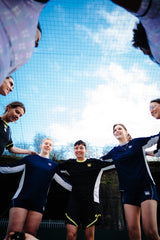 A group of girls and women wearing the Eleven as One blue training top, arms around each other.