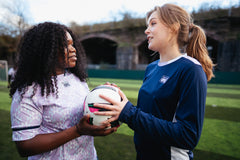 Two girls are chatting whilst holding a football. One is wearing the navy blue training top with long sleeves.