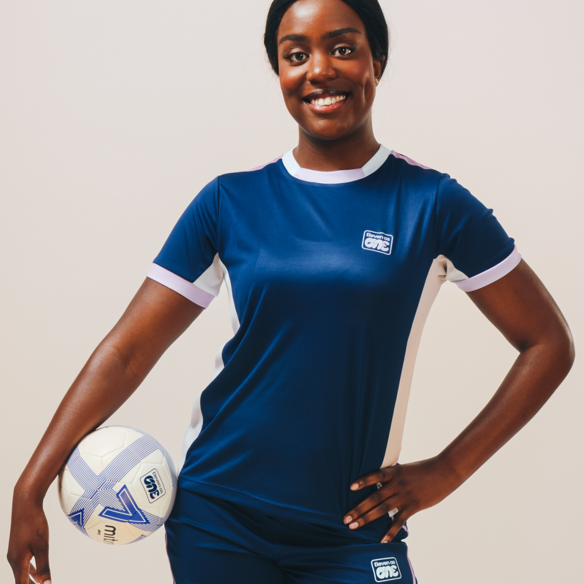 Girl stood with a hand on her hips and a football under the other arm wearing the Eleven as One blue round neck training top.