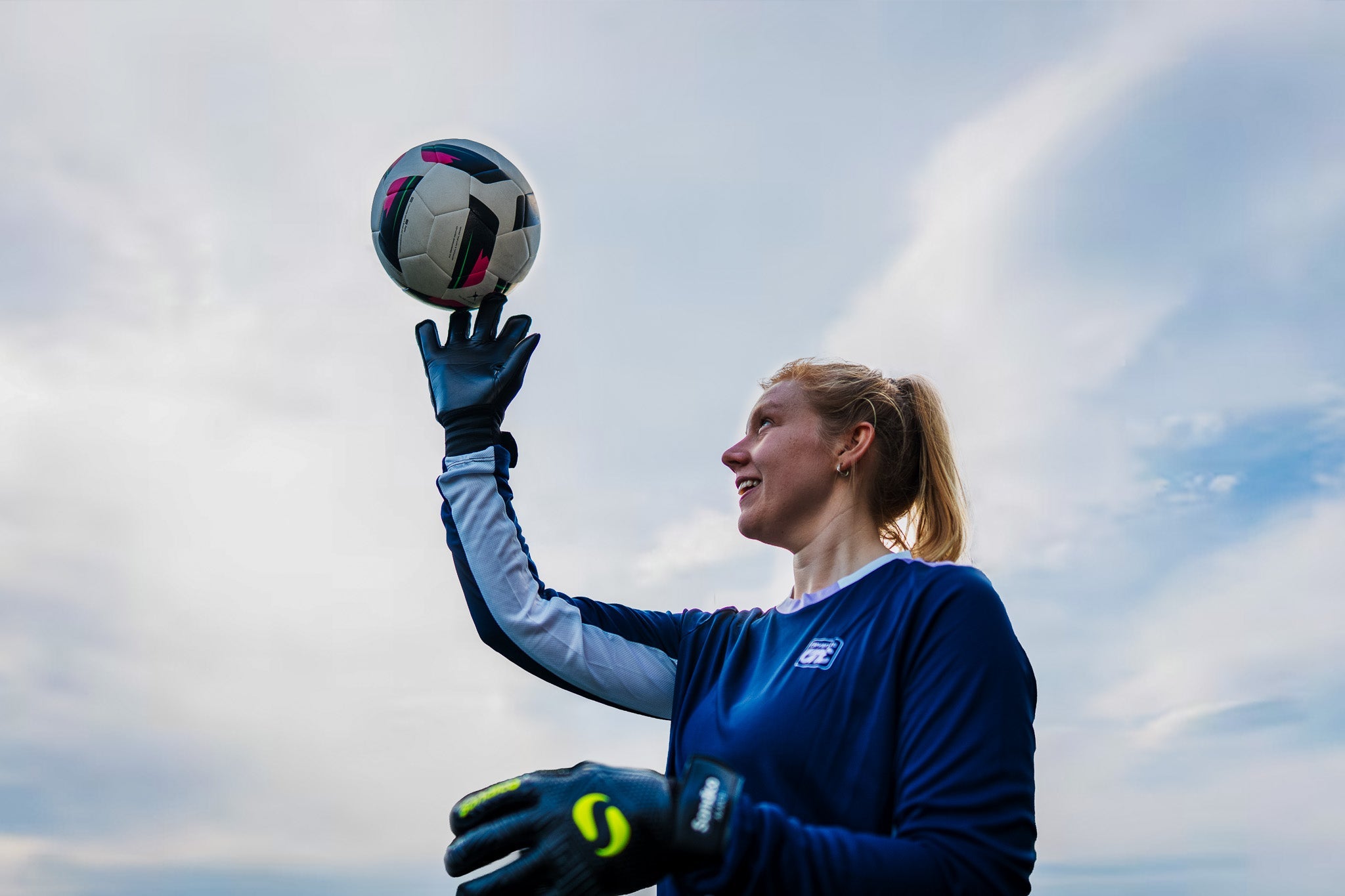 A girl with blond hair is wearing the navy blue training top with long sleeves, football gloves and is balancing a ball on her right hand. 